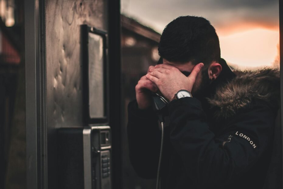 A man inside a phone booth appears worried, covering his face, during twilight.